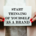 Person holding sign saying "start thinking of yourself as a brand"